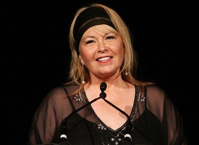 A smiling woman speaking at a microphone, wearing a black outfit and headband.